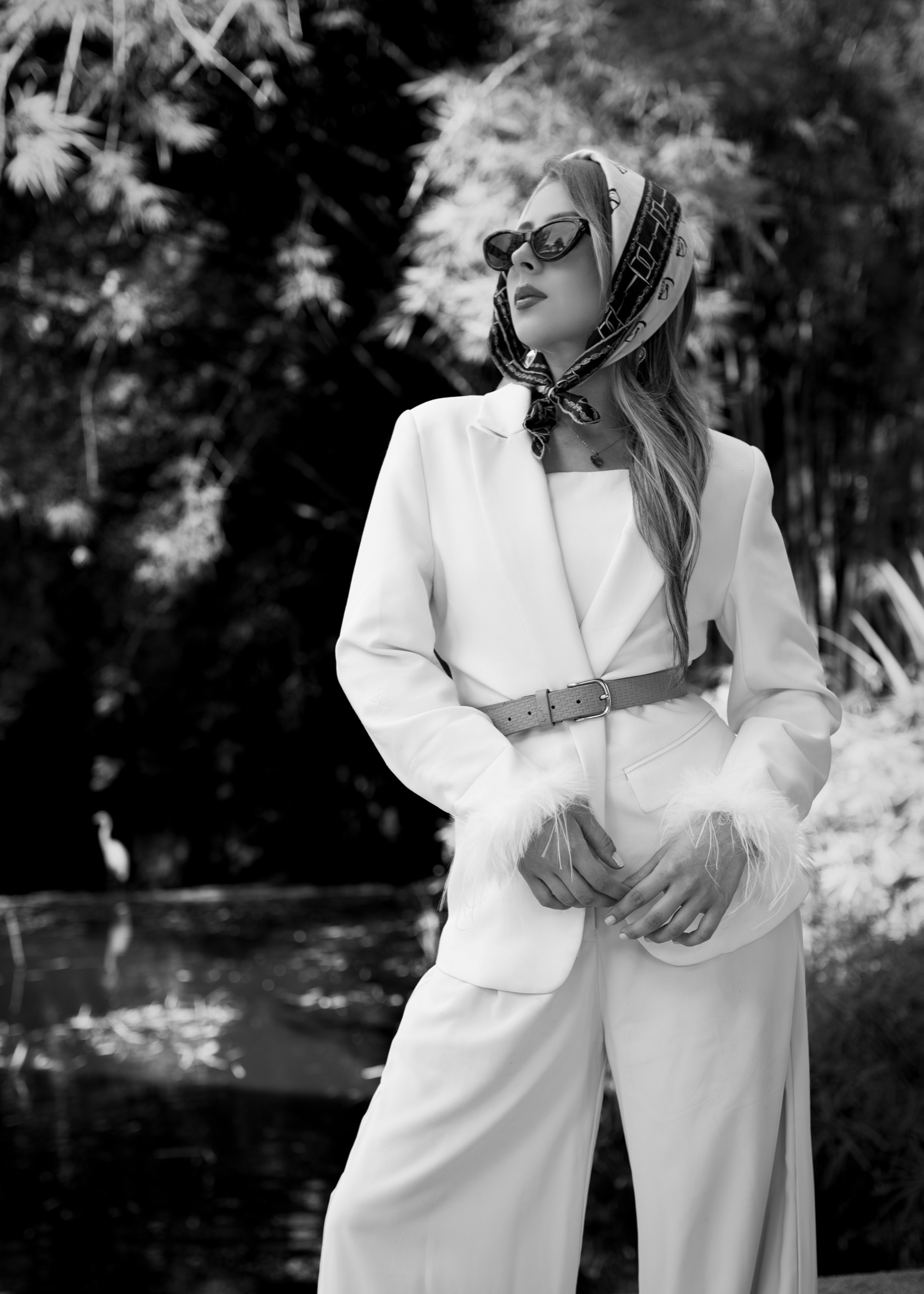 Modelo con traje blanco, pañoleta y gafas de sol junto a un lago, estilo editorial en blanco y negro