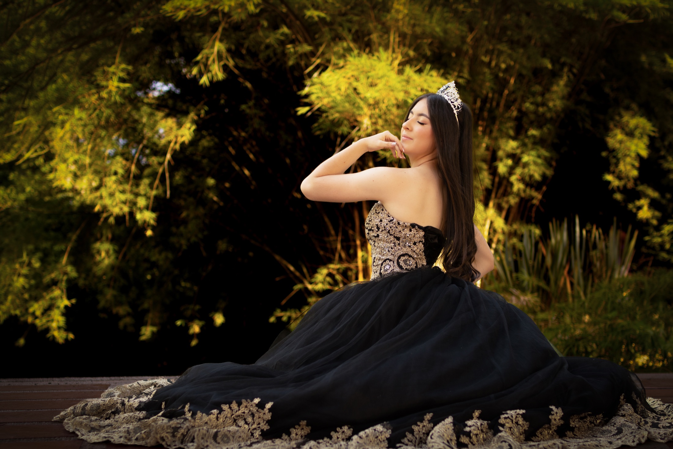 Quinceañera sentada con vestido negro y dorado en un jardín